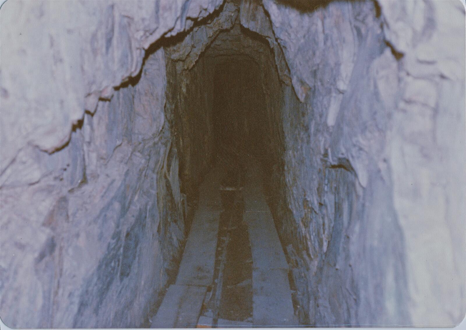 The interior of a narrow worked drift at the Red Wing in the 1980s, iron rails running down the floor, calcite-stained slate walls arching toward a low roof, receding into darkness.