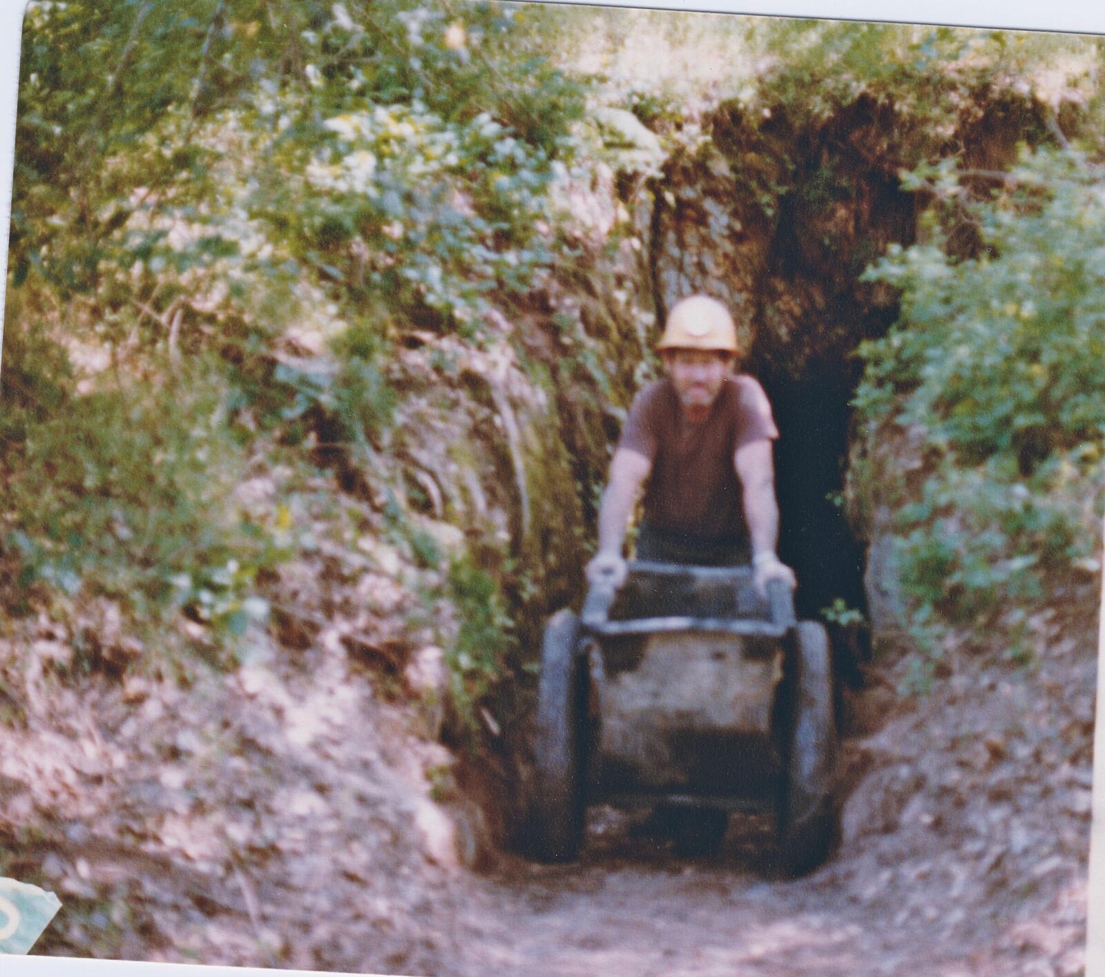 Gary pushing the loaded iron ore cart out of a wooden-framed portal entry at the Red Wing in the 1980s, red shirt, hard hat, gloves, hands on the rear of the cart.