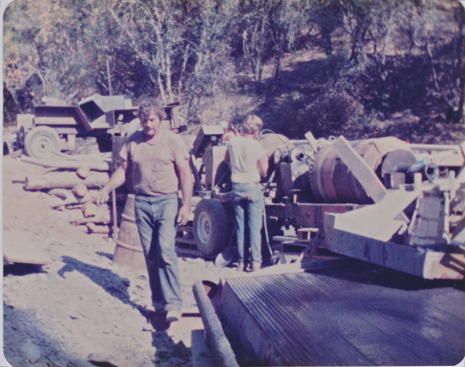 The Red Wing mill site in operation in the 1980s. Gary, Don's father, walks past the mill equipment; Don as a kid is at the mill drum behind him. Lumber stacked, rocker box in the foreground, oaks behind.