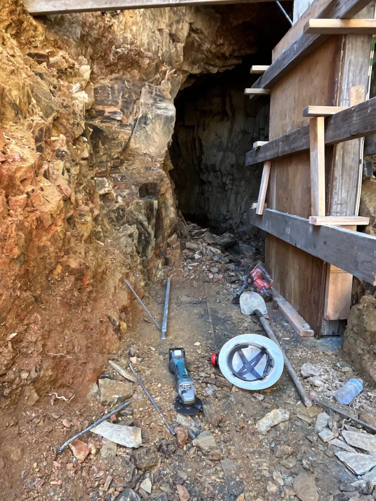 Looking down into the working pit during the portal-securing work, with a yellow hard hat, a Bosch rotor hammer, drill bits, rebar lengths, water bottle, and tape measure scattered around the half-built frame.