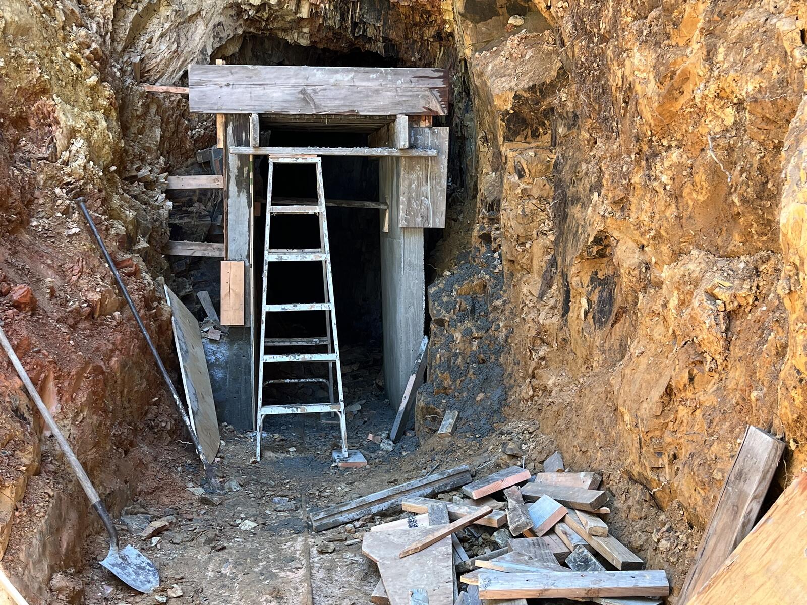 The completed wooden form for the concrete portal at the Red Wing, with an aluminum ladder going down into the cavity where the final pour will land, scrap timber stacked around the working face, and the rock face of the entry visible.