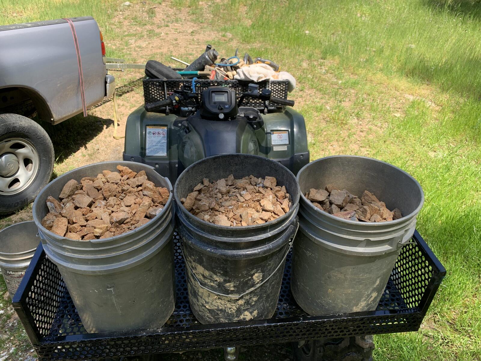 Three plastic buckets full of broken ore loaded onto the rack of Don's quad on grass at the staging area.