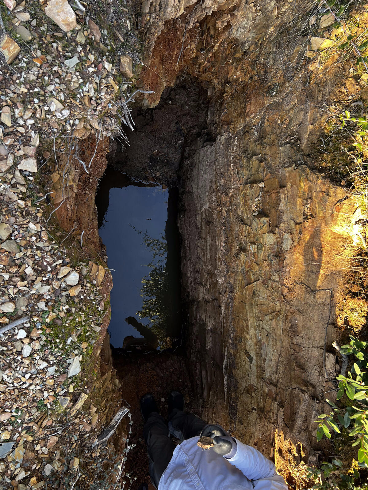 A surface trench on the Red Wing property near the top of the hill, looking down from the rim, with water standing in the bottom and a gloved hand visible at the foot of the cut.